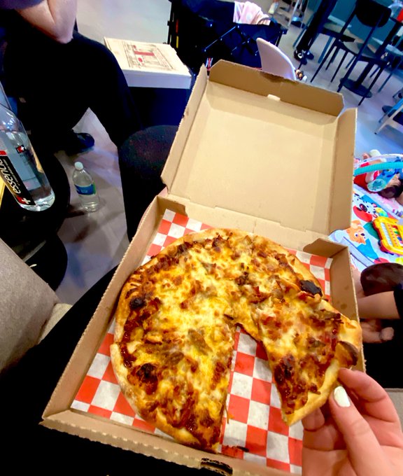A hand holds a slice of cheese pizza lifted from a delivery box lined with red and white checkered paper, enjoyed during a casual "Pizza watch party" as suggested by the post, with a water bottle and people seated in the background.