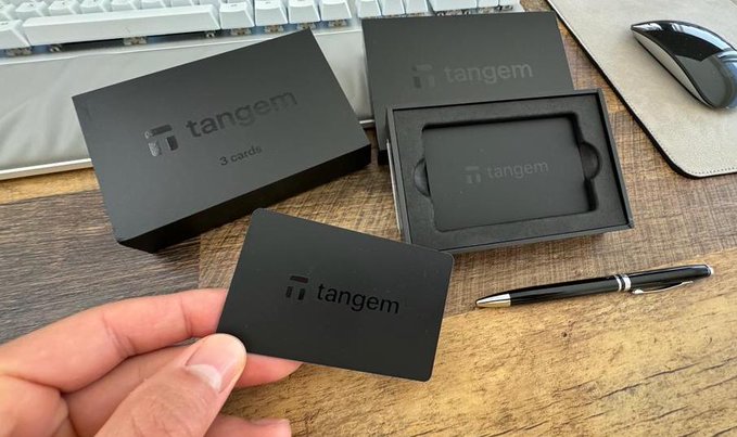 A hand holding a black 'Tangem' card, with packaging labeled 'Tangem 3 cards' and a card tray on a desk beside a keyboard, pen, and mouse.