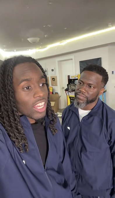 Two Black men wearing similar navy blue jackets stand indoors, possibly in a backstage area, with one looking off-camera and speaking.
