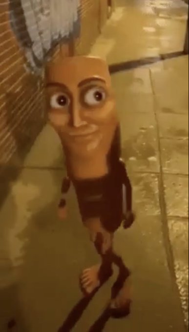 A bizarre CGI character with an elongated head, exaggerated facial features, and detailed tattoos stands on a sidewalk at night.