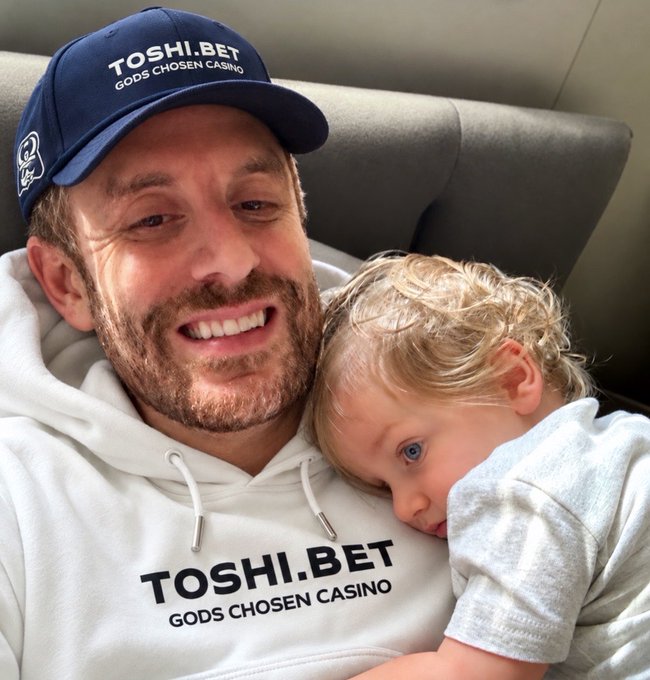 A smiling man wearing a navy 'TOSHI.BET GODS CHOSEN CASINO' cap and matching white hoodie cuddles a young blonde child resting against his chest.