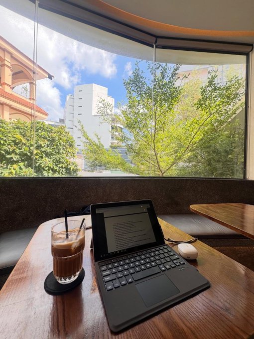 A cozy photograph taken from a café table showing an iced coffee in a glass, a tablet displaying text, and wireless earbuds, with a view of trees and city buildings through a large window.