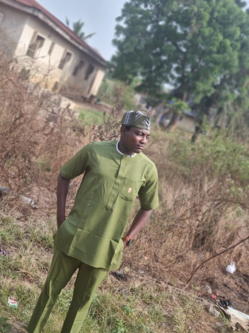 A man stands outdoors in a green traditional African outfit and patterned cap, posed in front of an overgrown area with an old building in the background.