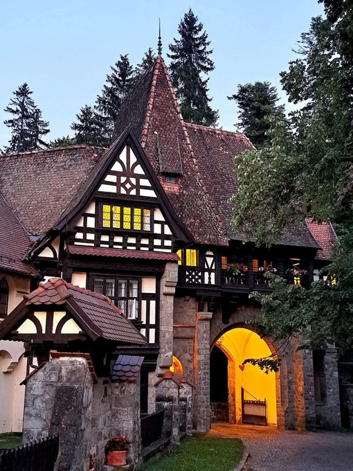 A picturesque view of a traditional Tudor-style building with a steep gabled roof, decorative timber framing, and an illuminated stone archway entrance at dusk.