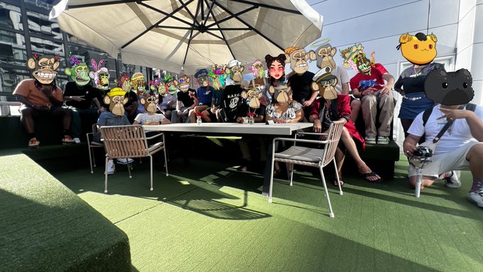 An outdoor group photo of people sitting on a tiered seating area under an umbrella. All faces in the group have been obscured by various drawn or pasted digital monster and character masks.