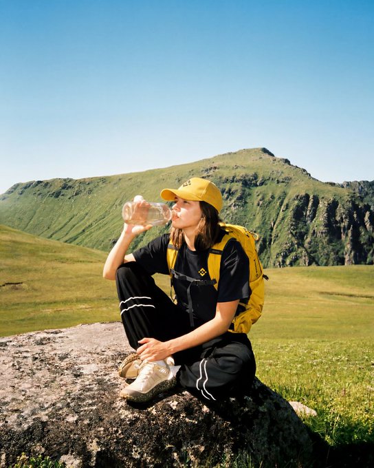 A hiker in a yellow cap and backpack drinks from a plastic bottle while resting on a rock amid green mountain ridges, embodying the spirit of "GM整装待发!" ("GM, ready to go!")—a fitting visual of being fully geared up and prepared for the journey ahead.