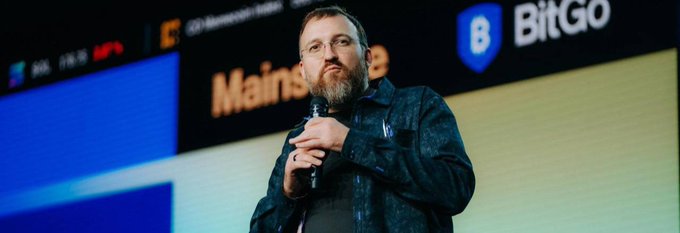 A bearded man in glasses and jacket speaks into a microphone on stage, with 'BitGo' branding and 'Mainstage' visible on the screen behind him.