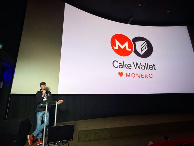 The image features a man standing behind a podium, speaking into a microphone during an event at a movie theater or auditorium. The main subject of his speech appears to be related to cryptocurrency, specifically Monero. The backdrop for this event includes a large screen displaying the words "Cake Wallet" and "Monero", indicating that it is likely a promotional event or conference about cryptocurrency.