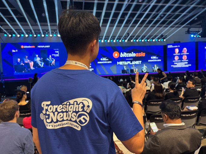 A person wearing a blue "Foresight News" t-shirt poses with a peace sign at the "bitcoin asia" and "METAPLANET" conference, standing in front of a stage with panelists and a large audience.