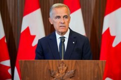 A man in a dark suit and tie stands at a wooden podium with Canadian flags displayed behind him.