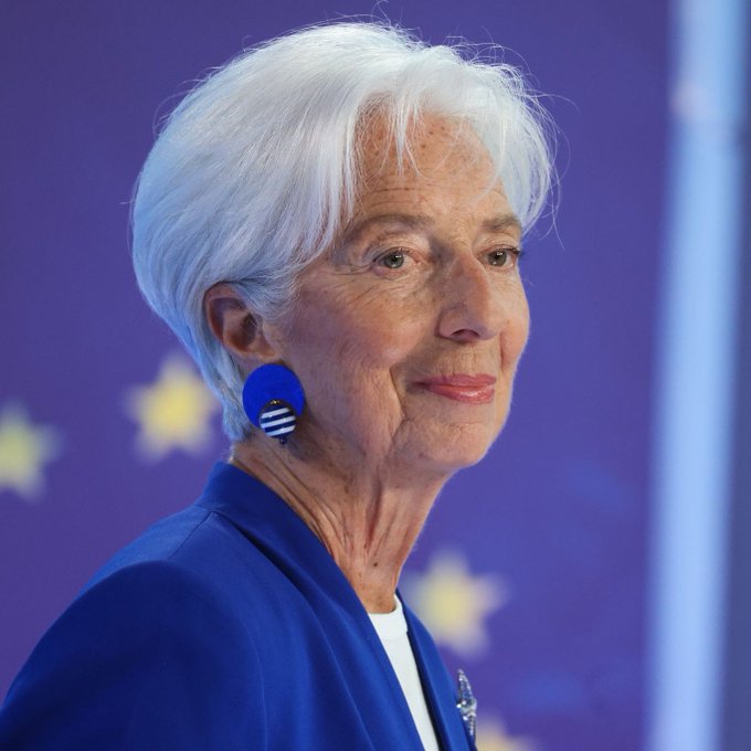 Christine Lagarde, President of the European Central Bank, photographed in profile with short white hair, wearing a blue blazer and distinctive blue earrings against a purple EU flag background.