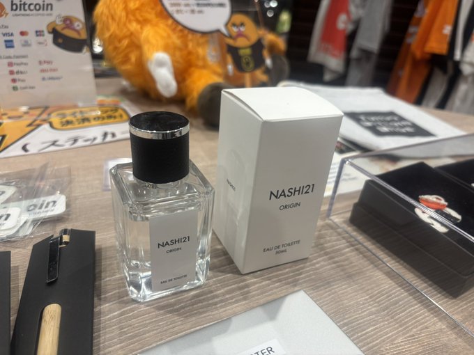 A bottle and box of 'NASHI 21 ORIGIN' Eau de Toilette for men, with a Bitcoin payment sign visible in the background.