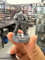 A hand holding up a small, painted miniature figure of a soldier or guard standing on a round base, photographed inside what appears to be a hobby store.