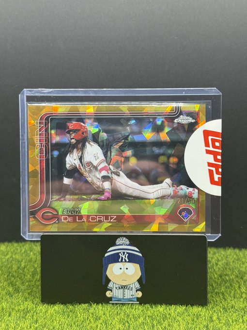 A Topps Chrome baseball trading card of Elly De La Cruz (Reds, #44/150) in a protective case, displayed next to a small South Park-style figurine wearing a Yankees hat.