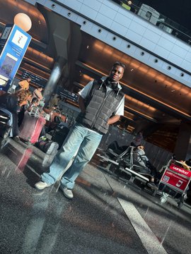 A man wearing a vest and light jeans poses confidently in what appears to be an airport terminal, with a red suitcase visible nearby, illustrating CryptoChris's lifestyle as a successful Nigerian trader celebrating his third house purchase in two years through Web3, crypto, and forex.