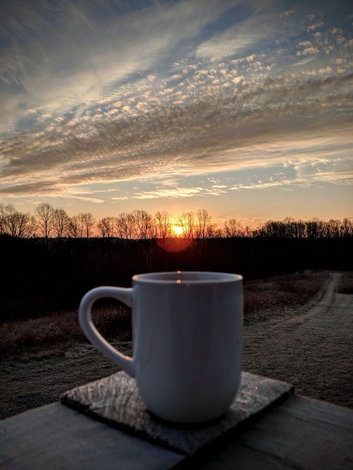 A white coffee mug sits on a wooden railing in the foreground, silhouetted against a dramatic sunrise or sunset with scattered clouds over a field.