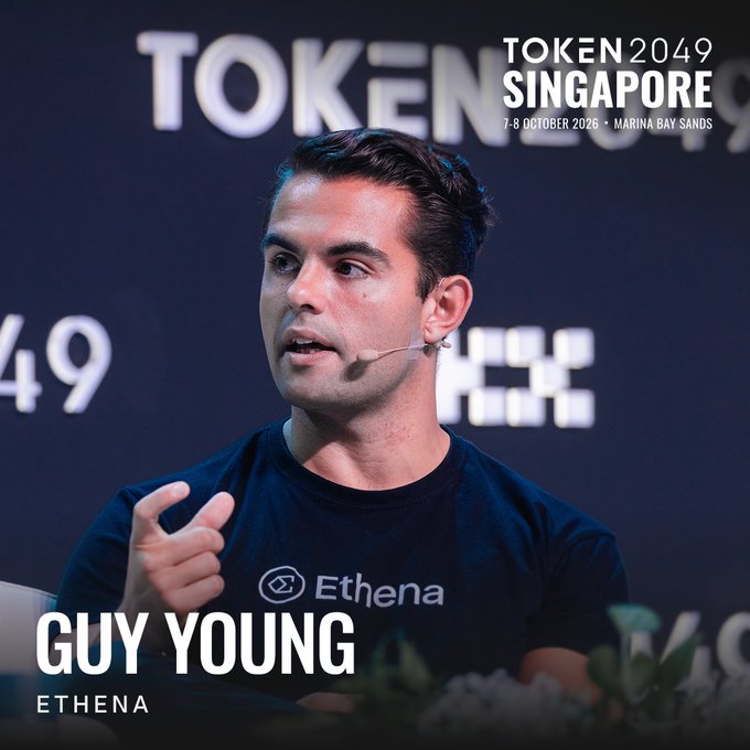 Guy Young, CEO of Ethena, is shown speaking at TOKEN2049 Singapore (October 7-8, 2026, Marina Bay Sands), wearing an Ethena-branded black t-shirt and headset microphone. The event branding in the background aligns with his featured appearance at the conference, highlighting Ethena's rapid growth from a $6M seed to a $15B stablecoin protocol.