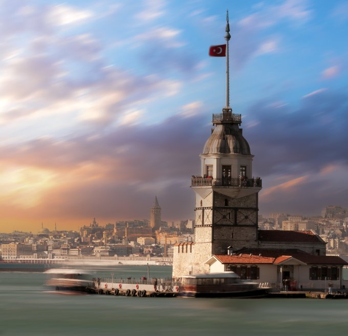 The Maiden's Tower (Kız Kulesi) in Istanbul, Turkey, standing on the Bosphorus with the Turkish flag flying on top, the Galata Tower visible in the background, and boats passing by.