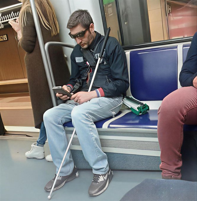 A visually impaired man wearing sunglasses sits on a subway seat while reading on a smartphone, accompanied by his white cane and a folded green cane, with other passengers visible nearby.