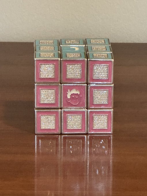 A decorative Rubik's cube-style puzzle with pink and teal faces featuring gold glitter accents, sitting on a wooden surface.