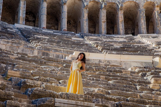 A woman in a yellow dress stands among the ancient stone tiers and arched columns of a Roman amphitheater. The image has no direct connection to the accompanying text about Binance careers and competitive job markets, appearing to be a generic travel photo unrelated to the message.