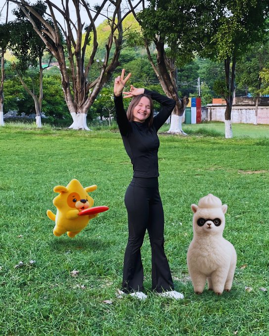 The image features a woman standing in a grassy field next to two stuffed animals - one dog and one llama. The woman appears to be posing for a photo, with her arms raised above her head as if she's celebrating or expressing joy. The background of the image is filled with trees, adding a natural element to the scene. The post includes text that reads "How good does it feel when the month starts on a Monday? Happy start of the week to the entire @Ronin_Network community! I hope you all have an amazing week, a fantastic start to the month, and, why not, a life filled with blessings and happy moments. I had a pretty". The woman's pose in front of the stuffed animals seems to be connected to this post, as if she is celebrating or expressing joy over her encounter with these cute toys.