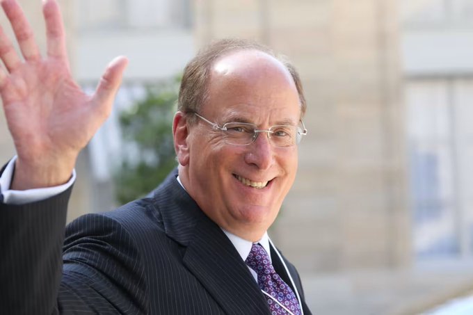 A smiling man in a dark pinstripe suit and purple tie waves at the camera while wearing rimless glasses, set against a blurred indoor background.