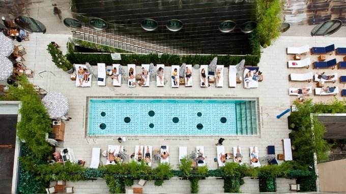 An aerial view of a rooftop pool deck at a hotel shows a rectangular swimming pool surrounded by sun loungers and people relaxing under umbrellas.