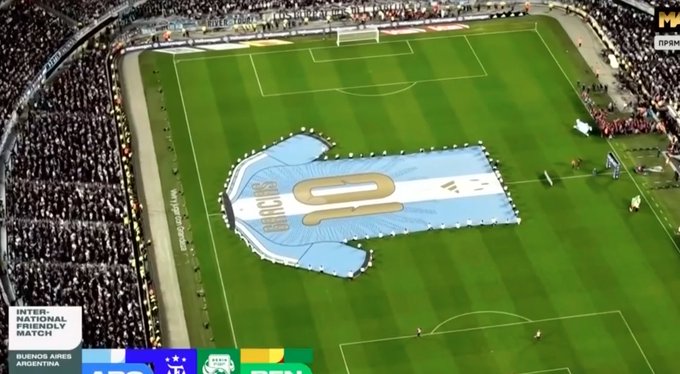 An aerial view of a soccer stadium where fans are holding up a giant light blue Argentina jersey with 'MESSI 10' printed on it during an international friendly match.