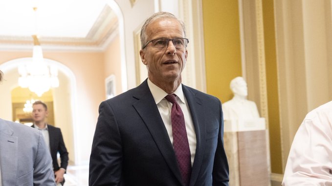 Senator John Thune, wearing a dark suit, white shirt, and maroon tie, walks through a brightly lit hallway with other people and a white bust visible in the background.