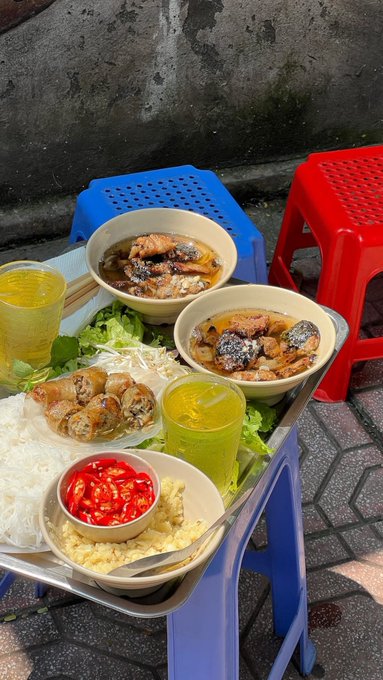 A street food meal featuring two bowls of soup with meat and mushrooms, spring rolls, rice noodles, fresh herbs, and chili, served on a blue plastic table with red stools.