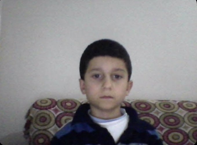A young boy with short dark hair sits on a patterned couch against a plain wall, wearing a striped sweater over a white shirt.