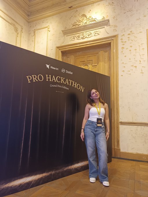 A woman in a white tank top, baggy blue jeans, and a yellow lanyard poses next to a large black backdrop reading "PRO HACKATHON · Grand Peru Edition" with Rise In and Stellar logos, inside an ornate room with decorative molding.