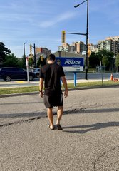 A person in a black t-shirt, black shorts, and a face mask walks away from the camera on a paved path near a police vehicle and street intersection.