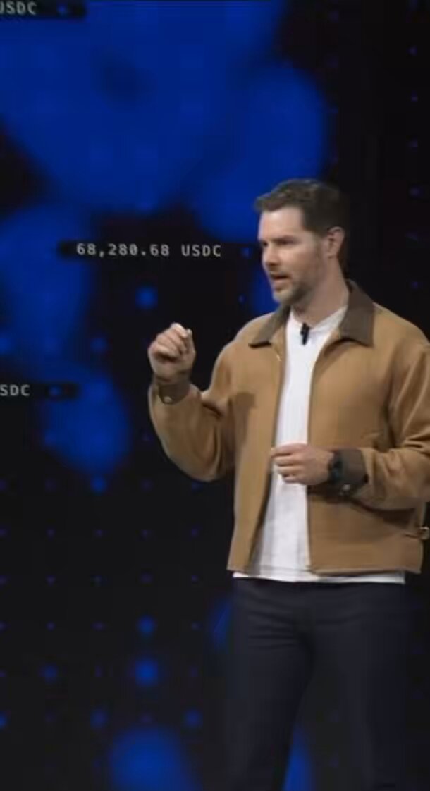 A man wearing a beige jacket stands in front of a large screen that displays some text, including "Best way to position yourself for $BASE on @base Base is backed by @coinbase ($574m raised) Second largest CEX Since the launched x402 last year  Over $185M in transactions ... 90% of onchain agentic stablecoin transaction volume running on @base".