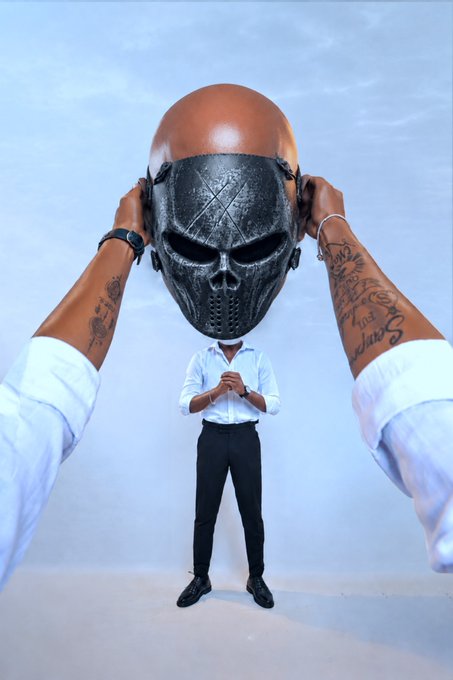 A surreal digital illustration shows a person in a white shirt and black pants standing with hands clasped, while giant tattooed arms hold a menacing dark skull mask above their head against a light blue background.