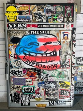 A door covered in numerous stickers, graffiti, and tags, with prominent text including 'VEKS', 'ZEE NIKE WERE HERE', 'STIGMATA', and 'THE SUA'.