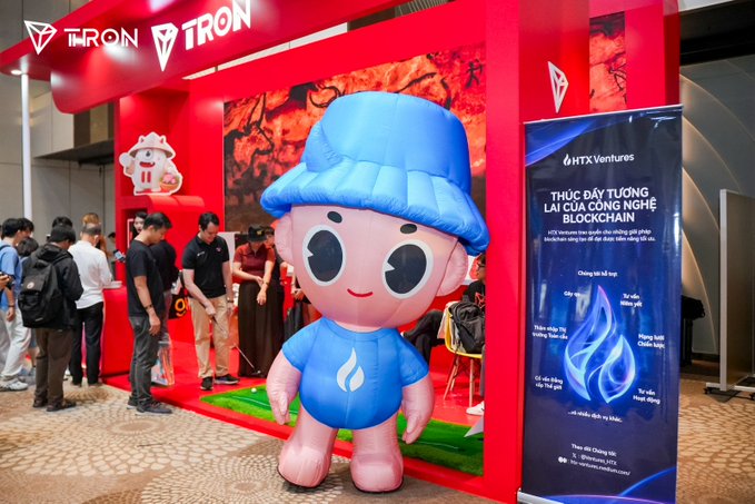 A large inflatable mascot wearing a blue cap and blue shirt stands in front of a TRON-branded booth at the Conviction 2026 event, with an HTX Ventures banner advertising blockchain training courses ("THÚC ĐẨY TƯƠNG LAI CỦA CÔNG NGHỆ BLOCKCHAIN") visible alongside attendees exploring the booth.