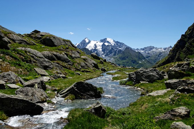 A bright alpine landscape with a rocky stream winding through green mountain slopes toward snow-capped peaks.