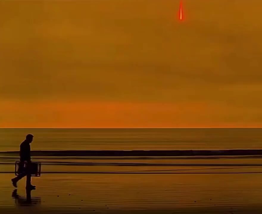 The image shows a man standing at the edge of a beach, with his suitcase nearby. He appears to be looking out towards the ocean or sky, possibly contemplating something. The photo has been edited to emphasize the contrast between the bright orange sky and the dark blue water below, creating an interesting visual effect. The caption "Me holding to zero instead of panic selling for a loss" suggests that there is no immediate danger or urgency in this scene, as if the man is not panicking but rather calmly observing his surroundings.