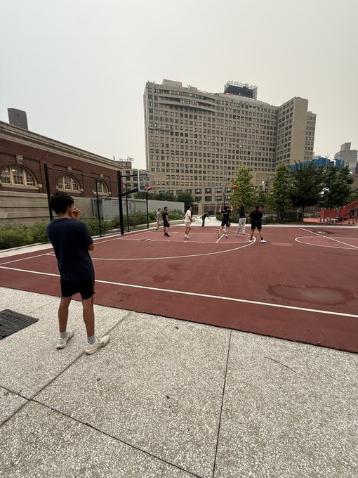 An outdoor basketball court scene where several people are playing or watching the game, with a tall residential building in the background.