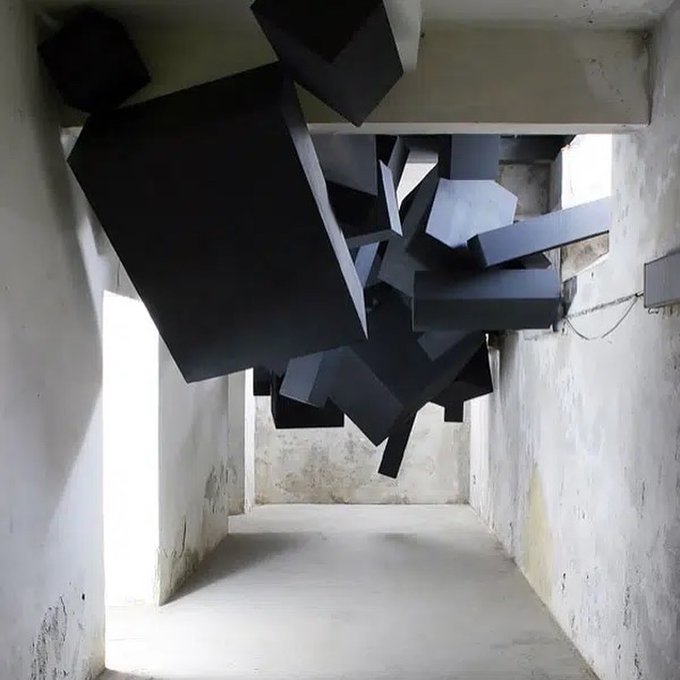 A cluster of black geometric blocks suspended from the ceiling in a dilapidated concrete room, creating an abstract sculpture installation.