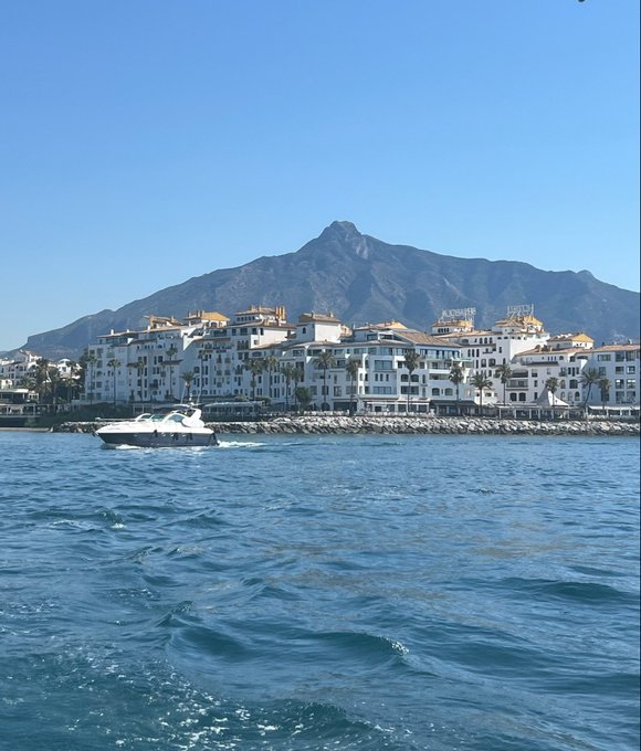 A scenic morning view of Puerto Banús along Spain's Costa del Sol, featuring whitewashed Mediterranean buildings along the waterfront with a prominent mountain in the background and a white yacht cruising the blue waters.