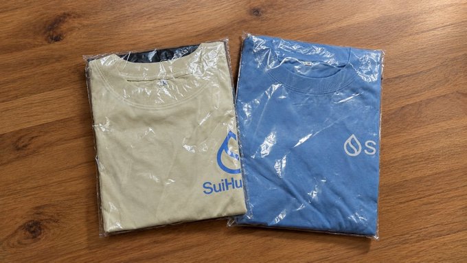 Two folded t-shirts in clear plastic packaging on a wooden surface; the beige one features 'SuiHu' branding while the blue one displays the Sui logo.
