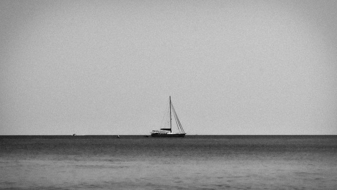 A black-and-white photograph of a lone sailboat on a calm sea beneath a pale sky, with subtle film grain visible across the image.