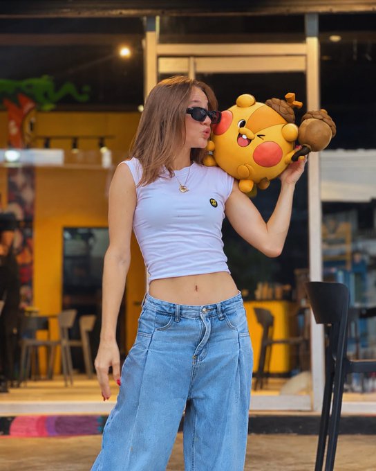 A young woman wearing sunglasses, a white crop top, and loose blue jeans holds a large yellow plush toy resembling a bee character near her face on a city street.