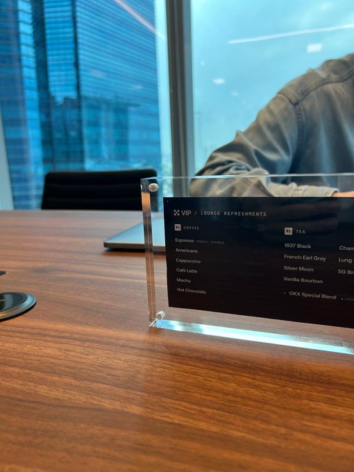A clear acrylic sign on a wooden table listing 'VIP LOUNGE REFRESHMENTS' with coffee options (Espresso, Americano, Cappuccino, Café Latte, Mocha, Hot Chocolate) and tea options (1837 Black, French Earl Grey, Silver Moon, Vanilla Bourbon, OKX Special Blend).