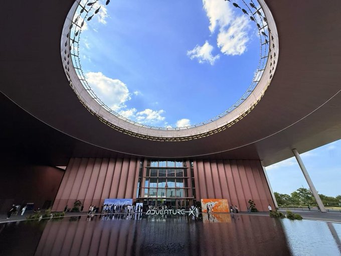 A large circular skylight opens to a bright blue sky above the entrance of the "AdventureX" venue, where visitors gather near a reflective water feature. The text "AdventureX" is visible on signage at the entryway, connecting the image to the post's mention of the AdventureX hackathon, where Injective emerged as a popular integration choice among diverse builders.