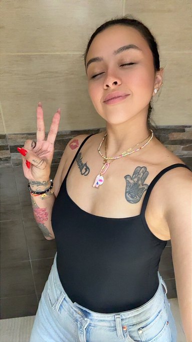 A young woman with tattoos on her chest and arm poses with a peace sign, wearing a black tank top and layered necklaces including one with a rainbow pendant.