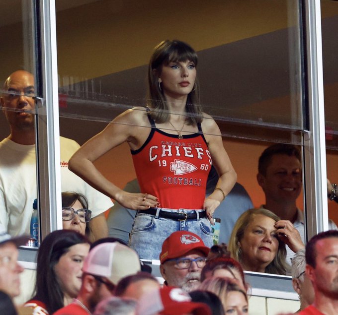 Taylor Swift stands in a luxury stadium suite wearing a red Kansas City Chiefs tank top with '19-60 FOOTBALL' text, surrounded by other spectators including a man in a red Chiefs cap.